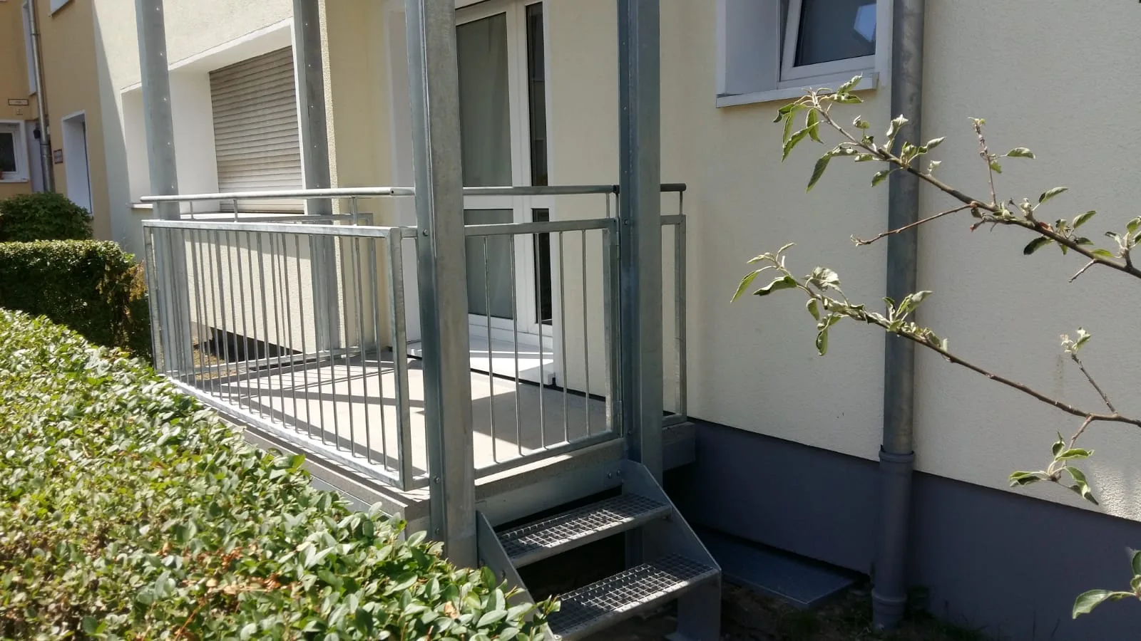 Feuerverzinkter Balkon im Erdgeschoss mit Stabgeländer und einer Treppe mit Gitterroststufen hinunter in den Garten.