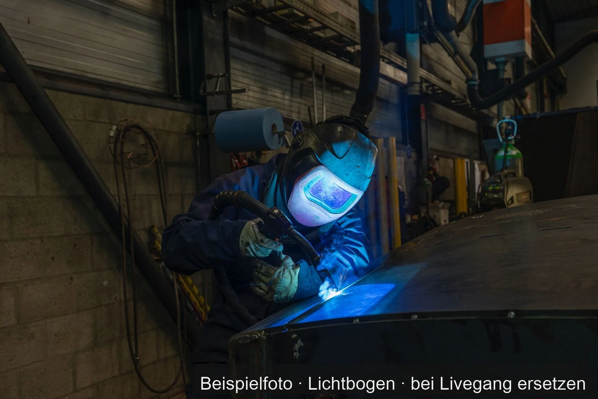 Ein Schweißer in dunkler Arbeitskleidung, mit Schweißhelm und Lederhandschuhen, schweißt an einer großen, gewölbten Blechkonstruktion. Dahinter eine Werkstattwand aus Kalksandstein mit Regalen, Kabeln und einer Absauganlage.