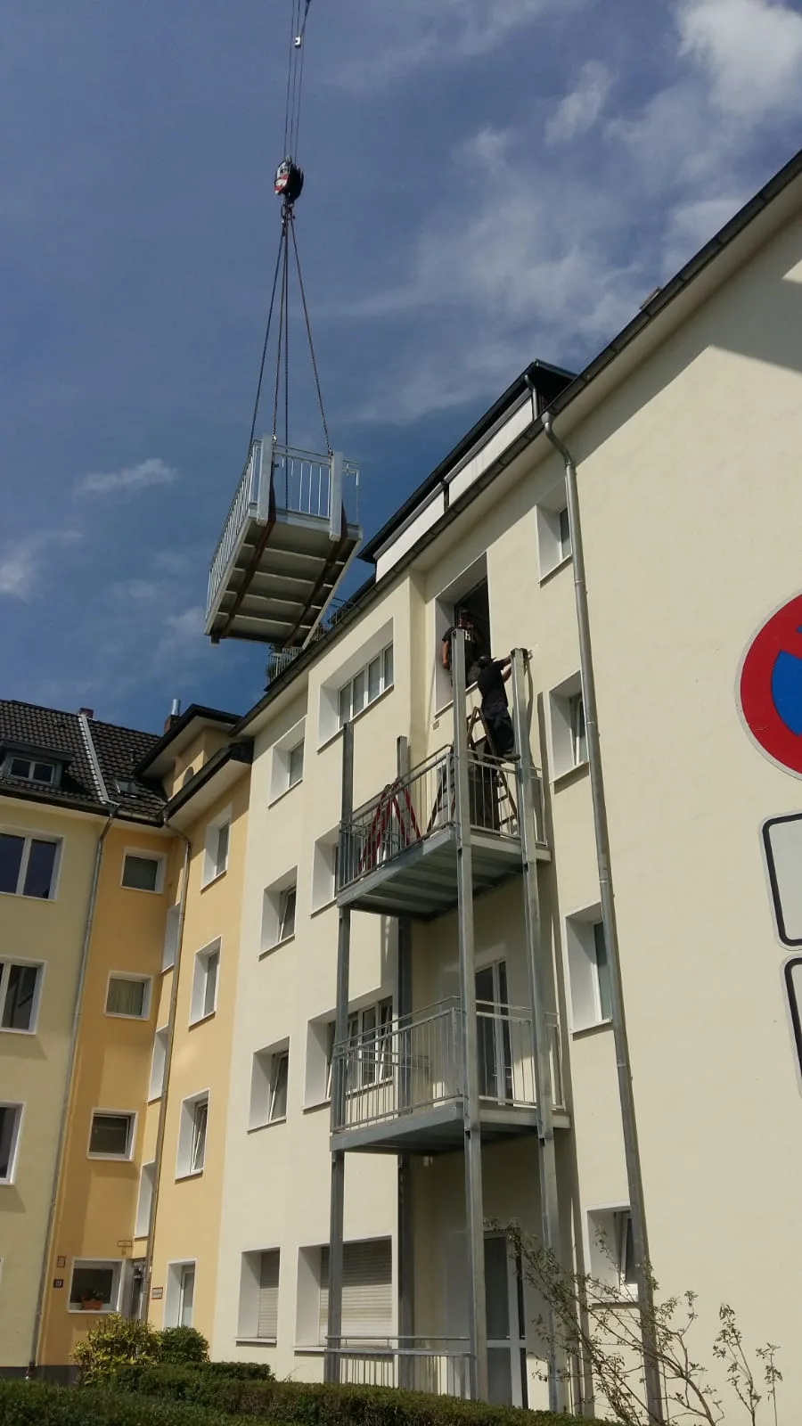Ein Autokran hebt ein Balkonelement an ein mehrgeschossiges Wohnhaus, ein Monteur nimmt es am Balkon in Empfang.